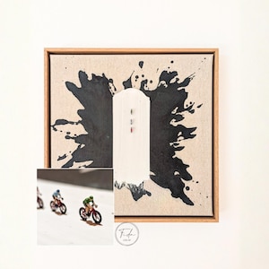 Könnte beinhalten: Abstraktes Kunstwerk mit einem schwarzen Tintenspritzer-Design auf einer beigefarbenen Leinwand, mit einer weißen vertikalen Form in der Mitte. Ein kleiner Einsatz zeigt Miniatur-Radfahrer auf Fahrrädern. Das Kunstwerk ist in einem Naturholzrahmen gerahmt.