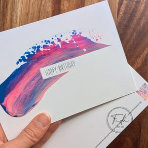 Può includere: Biglietto di auguri bianco con un design astratto a pennellate blu e rosa. Le parole "HAPPY BIRTHDAY" sono stampate su un rettangolo bianco. Il biglietto è tenuto in mano.