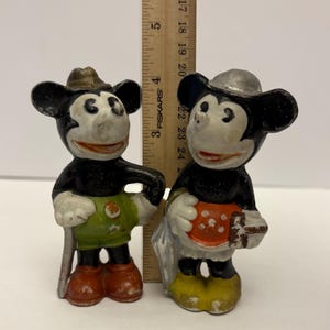 May include: Two vintage Mickey Mouse figurines. One Mickey Mouse figure is wearing a brown hat, green shorts, and red shoes. The other Mickey Mouse figure is wearing a silver hat, an orange apron with white polka dots, and yellow shoes.