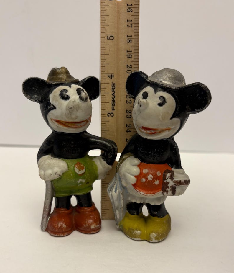May include: Two vintage Mickey Mouse figurines. One Mickey Mouse figure is wearing a brown hat, green shorts, and red shoes. The other Mickey Mouse figure is wearing a silver hat, an orange apron with white polka dots, and yellow shoes.