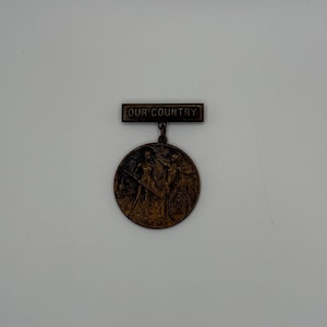 May include: A bronze-toned medal with a rectangular bar above it. The bar reads "OUR COUNTRY" in capital letters. The circular medal features a detailed relief of figures and text.