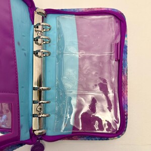 May include: Open, zippered binder with a colorful, galaxy-like pattern. The interior features a purple and blue color scheme, with a silver, three-ring binder mechanism. Clear plastic pockets are visible, along with a small, clear window pocket.