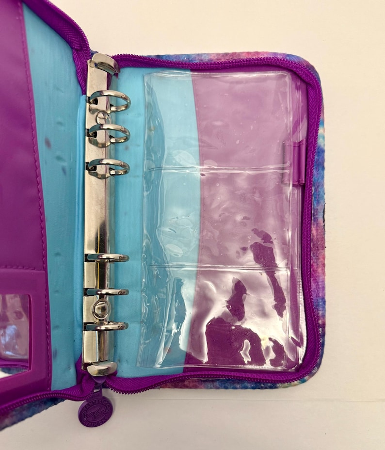 May include: Open, zippered binder with a colorful, galaxy-like pattern. The interior features a purple and blue color scheme, with a silver, three-ring binder mechanism. Clear plastic pockets are visible, along with a small, clear window pocket.