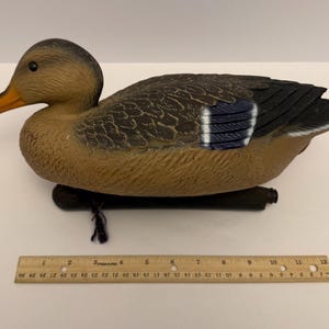 Puede incluir: Un señuelo de pato marrón y beige con una base negra y pico naranja. El pato tiene patrones de plumas detallados y detalles azules en sus alas. Una regla de madera se coloca debajo.