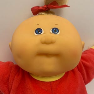 May include: A Cabbage Patch Kid doll with blonde hair tied up in a ponytail with a red bow. The doll is wearing a red and yellow sleeper.