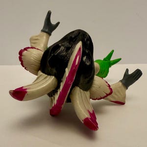 May include: A unique, abstract figurine with a black head and cream-colored appendages with pink and red accents. The figure has two arms with gray gloves and a green element. The design is artistic and eye-catching.