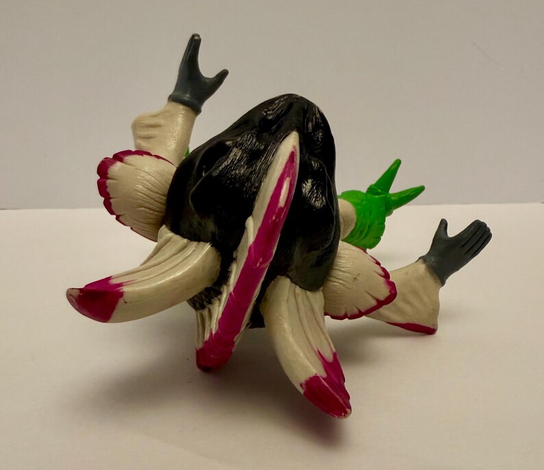 May include: A unique, abstract figurine with a black head and cream-colored appendages with pink and red accents. The figure has two arms with gray gloves and a green element. The design is artistic and eye-catching.