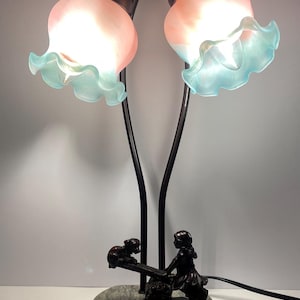 Op de afbeelding: Een zwarte metalen tafellamp met twee roze en blauwe glazen lampenkappen. De voet heeft een gesculpteerd tafereel van twee kinderen op een wip.