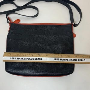 May include: Black crossbody bag with a red trim and strap. The bag is approximately 12 inches wide. The bag has a textured surface and a black strap. The text "LEES MARKETPLACE DEALS" is printed on a ruler.