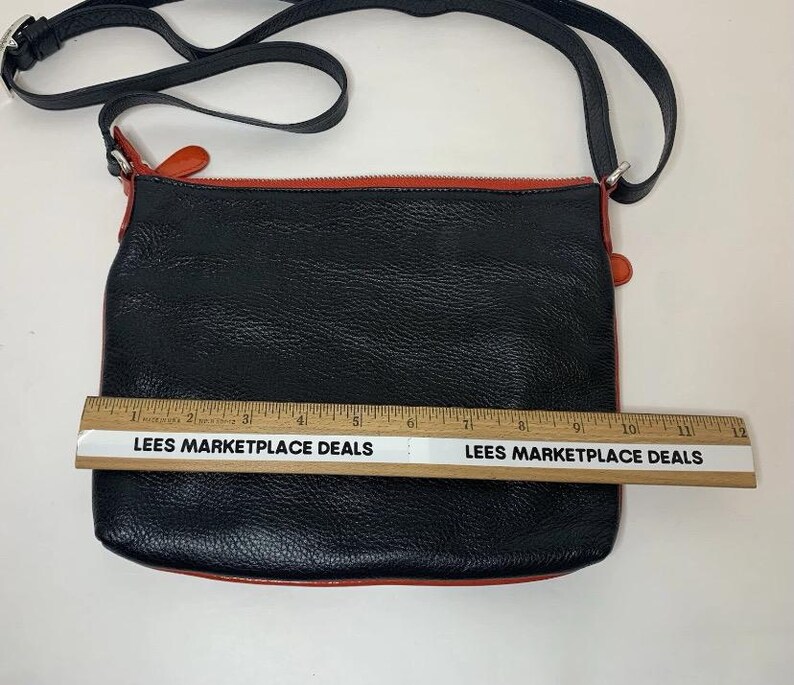 May include: Black crossbody bag with a red trim and strap. The bag is approximately 12 inches wide. The bag has a textured surface and a black strap. The text "LEES MARKETPLACE DEALS" is printed on a ruler.