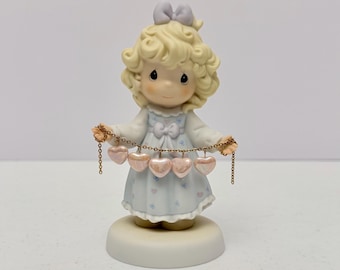 Może przedstawiać: Porcelanowa figurka dziecka z kręconymi blond włosami i jasnofioletową kokardą, ubranego w jasnoniebieską sukienkę w drobny kwiatowy wzór. Figurka trzyma złoty metalowy łańcuszek z pięcioma błyszczącymi, jasnoróżowymi zawieszkami w kształcie serc.