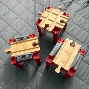 May include: Three wooden toy train track pieces with red plastic support legs. One piece is a four-way intersection, while the other two are straight track segments featuring grey plastic side rails. The tracks have a natural wood grain finish.