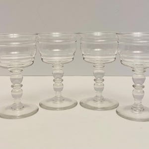 May include: Four clear, stemmed glasses with a decorative, horizontal-ridged design on the bowl. The stems have a series of rounded features, and the glasses sit on a round base. The glasses are arranged in a row.