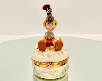 May include: A porcelain trinket box featuring a figurine of Pinocchio sitting on the lid with Jiminy Cricket perched on his head. The round base is decorated with small pink and blue flowers and has a gold-toned metal hinge and clasp closure.