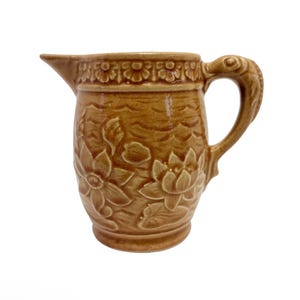 Könnte beinhalten: Braune Keramikkrug mit floralem Design. Der Krug hat einen Ausguss und einen Griff mit einem vogelartigen Design. Der Körper des Kruges ist mit geprägten Blumen und Blättern verziert. Die Oberseite des Kruges hat einen dekorativen Rand.