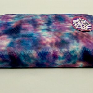 May include: A rectangular, zippered pouch with a tie-dye pattern in shades of blue, purple, and white. A circular patch with text is affixed to the top. The zipper pull is a matching purple color.