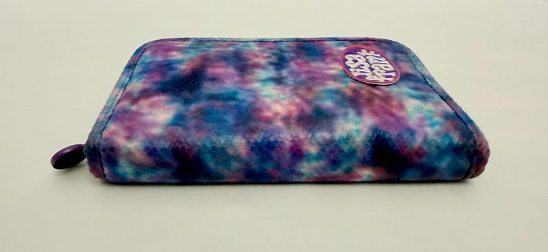 May include: A rectangular, zippered pouch with a tie-dye pattern in shades of blue, purple, and white. A circular patch with text is affixed to the top. The zipper pull is a matching purple color.