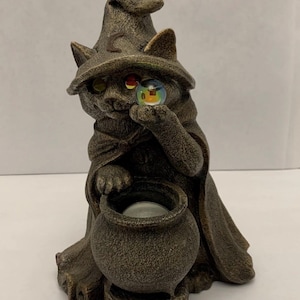 May include: A bronze-colored cat figurine wearing a witch's hat and cloak. The cat holds a crystal ball in one paw and sits next to a cauldron. The figurine has a detailed, textured finish, perfect for Halloween decor.