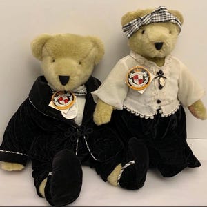 Può includere: Due orsacchiotti di peluche vestiti con abiti formali. Uno indossa un abito di velluto nero con finiture bianche, l'altro una camicetta bianca, una gonna nera e una fascia a scacchi. Entrambi hanno etichette con uno stemma.