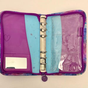 May include: Open, zippered binder with a colorful, tie-dye-like pattern. The interior features purple and light blue sections, a mirror, and clear pockets. A silver metal ring binder mechanism is visible.