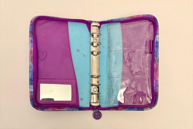 May include: Open, zippered binder with a colorful, tie-dye-like pattern. The interior features purple and light blue sections, a mirror, and clear pockets. A silver metal ring binder mechanism is visible.