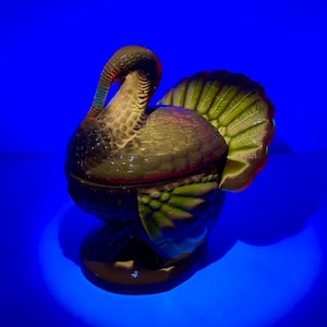 Op de afbeelding: Een keramieken kalkoenfiguur met een donker lichaam, groene staartveren en een gebogen nek. De kalkoen staat op een ronde basis. De kleuren zijn donkergroen, bruin en rood. De achtergrond is helderblauw.