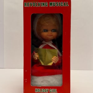 May include: A vintage Christmas decoration featuring a doll in a red dress and white fur-trimmed hood, holding a golden book. The doll is inside a red box with the text "REVOLVING MUSICAL" and "HOLIDAY GIRL DECORATION".
