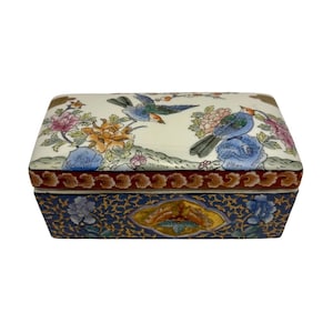 May include: Rectangular ceramic trinket box with a cream-colored lid featuring colorful birds and floral designs. The box's base is navy blue with gold accents and floral patterns. The box has a red border.