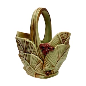 May include: A ceramic basket-style vase with a handle, featuring a leaf design in shades of green and brown. The vase has a decorative cluster of red berries. The overall design is reminiscent of nature.