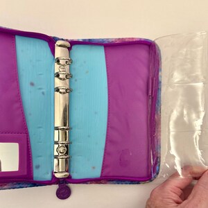 May include: Open purple and blue binder with silver rings and a clear plastic sleeve. The binder has a zippered closure and a small mirror. The interior features a light blue section and a purple section.