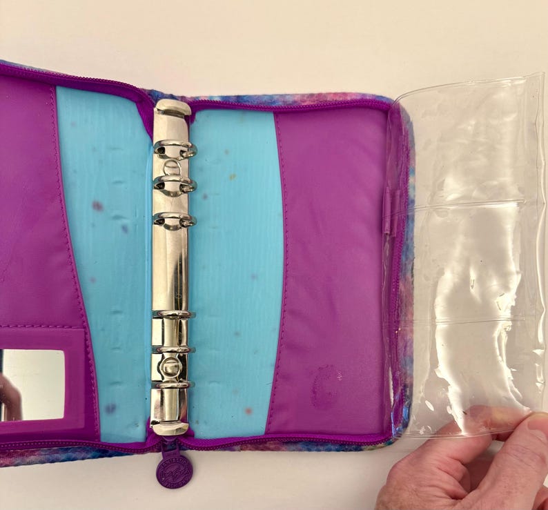 May include: Open purple and blue binder with silver rings and a clear plastic sleeve. The binder has a zippered closure and a small mirror. The interior features a light blue section and a purple section.