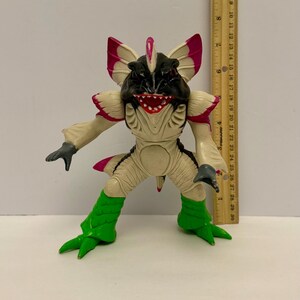 May include: A detailed action figure of a creature with a black head, cream-colored body, and green legs. The figure has pink accents on its ears and back. A ruler is next to the figure, showing measurements in inches.
