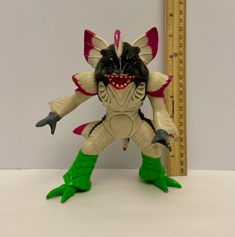 May include: A detailed action figure of a creature with a black head, cream-colored body, and green legs. The figure has pink accents on its ears and back. A ruler is next to the figure, showing measurements in inches.