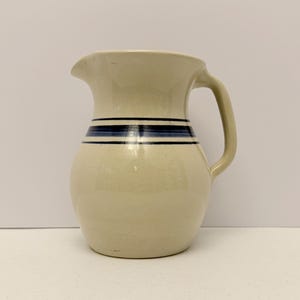 Puede incluir: Una jarra de cerámica de color crema con cuerpo redondeado, pico ancho y un asa lateral resistente. El exterior presenta una banda decorativa de tres rayas horizontales de color azul oscuro que rodean la parte superior del cuello.