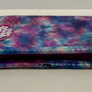 May include: A rectangular, tie-dye patterned wallet in shades of blue, purple, and pink. The wallet has a zipper closure and a small, circular patch with text. The overall design is a vibrant mix of colors.
