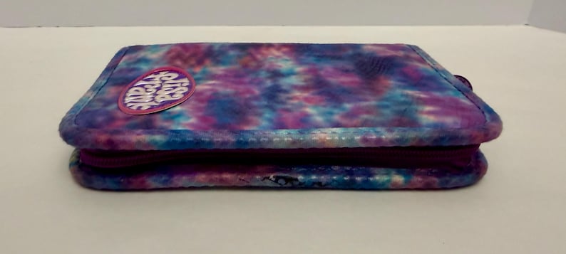 May include: A rectangular, tie-dye patterned wallet in shades of blue, purple, and pink. The wallet has a zipper closure and a small, circular patch with text. The overall design is a vibrant mix of colors.