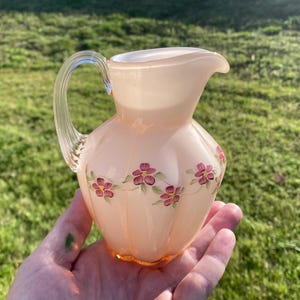 May include: A small, pale pink glass pitcher with a delicate floral design. The pitcher has a rounded body and a short, curved spout. The flowers are painted in shades of pink and green.
