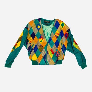 Peut inclure: Une veste en cuir à manches longues avec un motif losange multicolore dans des tons de bleu sarcelle, jaune, bleu et beige. La veste présente des symboles de cartes à jouer, un ourlet et des poignets côtelés sarcelle, une fermeture à pression et une doublure en satin sarcelle.