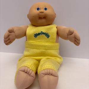 May include: A Cabbage Patch Kids doll with a bald head, blue eyes, and a small smile. The doll is wearing a yellow knitted outfit with the Cabbage Patch Kids logo on the front.