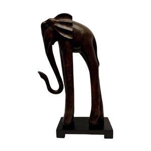 Op de afbeelding: Een donkerbruin houten olifant sculptuur met een gebogen slurf en lange poten. De olifant staat op een zwarte rechthoekige basis. Het beeldhouwwerk heeft een gladde, gepolijste afwerking, tegen een witte achtergrond.