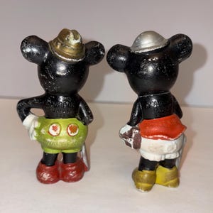 May include: Two vintage Mickey Mouse figurines. One wears a gold-colored hat, green shorts, red shoes, and white gloves. The other wears a silver hat, red skirt, yellow shoes, and white gloves. Both are black with white accents.