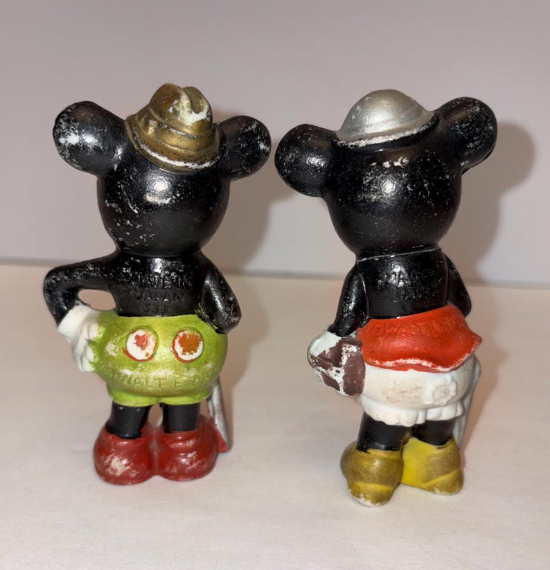 May include: Two vintage Mickey Mouse figurines. One wears a gold-colored hat, green shorts, red shoes, and white gloves. The other wears a silver hat, red skirt, yellow shoes, and white gloves. Both are black with white accents.