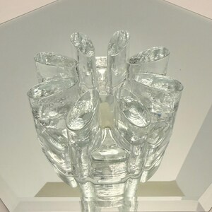Puede incluir: Un portavelas de cristal transparente con un diseño único. El portavelas presenta una columna central rodeada de ocho copas cilíndricas, creando una forma de flor. El cristal tiene una superficie texturizada, que refleja la luz.