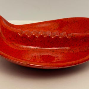 Könnte beinhalten: Ein leuchtend roter, nierenförmiger Aschenbecher aus Keramik mit glänzender Oberfläche und dunklen Sprenkeln. Das Stück hat ein geschwungenes, längliches Design mit einer Reihe von sechs integrierten Kerben am Innenrand zum sicheren Ablegen.