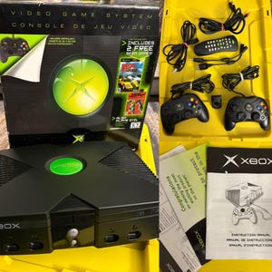 Puede incluir: Una consola de videojuegos Xbox negra con un logotipo circular verde, dos mandos negros, un mando a distancia, cables de alimentación y AV, y un manual de instrucciones. La caja original incluye el texto Video Game System y dos juegos de Sega.