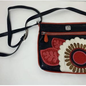 May include: Black crossbody bag with a decorative floral design in red, white, and brown. The bag has a black strap, red trim, and a silver zipper. The bag is made of leather.