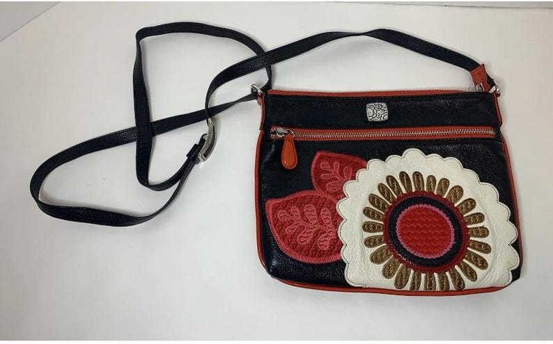 May include: Black crossbody bag with a decorative floral design in red, white, and brown. The bag has a black strap, red trim, and a silver zipper. The bag is made of leather.
