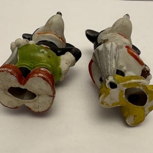 May include: Two vintage Mickey Mouse figurines, lying on their backs. The figures are painted with black, white, red, yellow, and green. One figure wears green shorts and red shoes. The other figure wears yellow shoes and a red and white outfit.