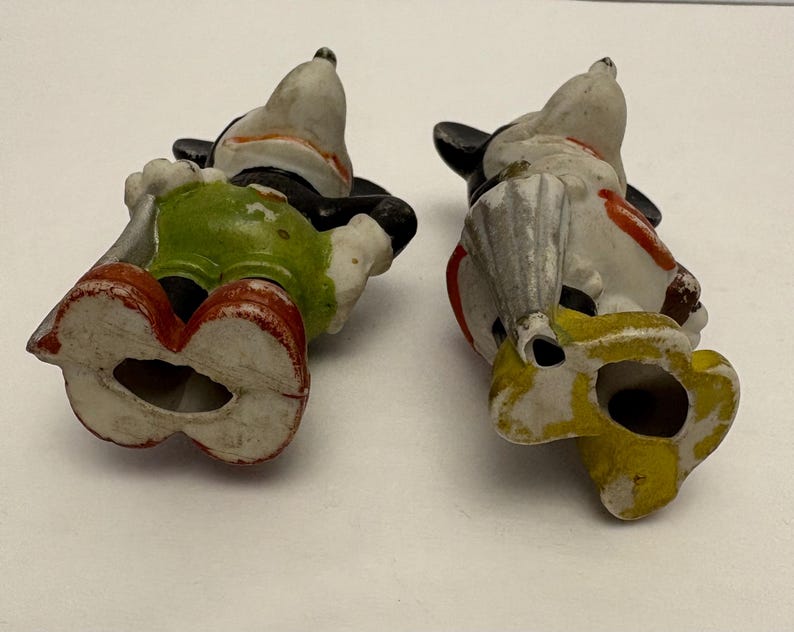 May include: Two vintage Mickey Mouse figurines, lying on their backs. The figures are painted with black, white, red, yellow, and green. One figure wears green shorts and red shoes. The other figure wears yellow shoes and a red and white outfit.