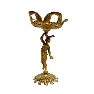May include: A gold-colored decorative statue featuring a classical figure holding a decorative bowl aloft. The statue has a detailed floral base and a detailed bowl with a leaf-like design. The figure is in a standing pose.